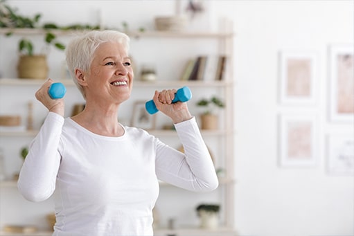Eine ältere Dame mit kurzem grauem Haar und weißem Oberteil lächelt, während sie eine Physiotherapie Übung mit kleinen Hanteln durchführt.