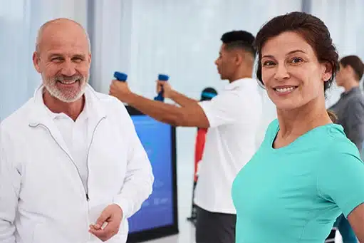 Pixformance-Selbstzahlerbereich-aufbauen-Physiotherapie-Unterstuetzung-Entlastung-Blog Zwei Physiotherapeuten im Vordergrund, lächelnd und froh über die Physiotherapie Unterstützung. Rechts eine Frau mit dunklem Haar und türkisem Shirt, links ein älterer Mann mit wenig Haar und weißer Jacke