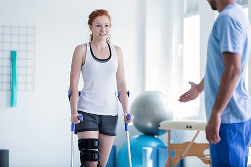 blog-bewegungsaktivierung-pixformance-rehabilitation-bewegungsanalyse-patienten-aktivieren-physiotherapie-6 Junge Patientin mit einer Knieverletzung grinsend auf ihre Krücken gestützt mit einem Physiotherapeuten unscharf im Vordergrund, der ihr mithilfe von Gesten etwas erklärt