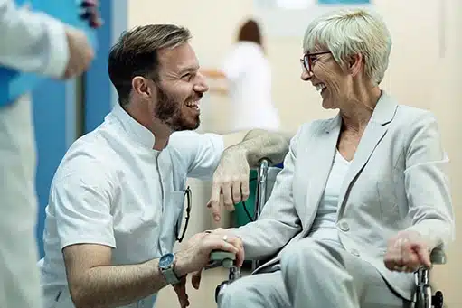 blog-bewegungsaktivierung-pixformance-rehabilitation-bewegungsanalyse-patienten-aktivieren-physiotherapie-7 Junger Arzt in der Hocke neben einer älteren Patientin im Rollstuhl. Beide sehen sehr glücklich aus und strahlen.