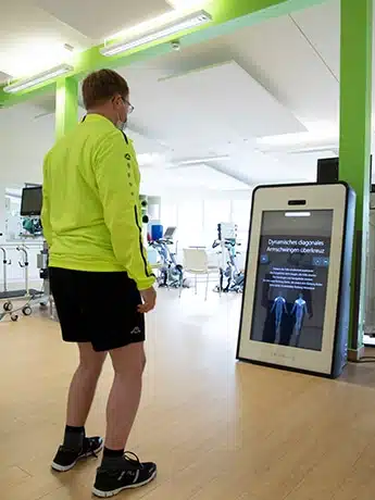 Pixformance-Sauerland-Klinik-Hachen-Telerehabilitation-Therapie-3 Telerehabilitation in action at the Sauerlandklinik Hachen: A patient wearing a yellow jacket and black shorts exercises in front of the Pixformance ward